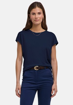 Vrouw draagt een donkerblauw mouwloos topje gestopt in een donkerblauwe broek met een bruine riem, gouden creoolhangers en een dunne gouden ketting.