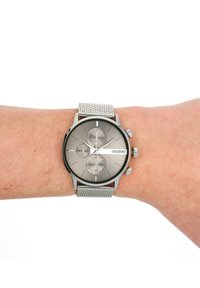 Montre en acier inoxydable argentée avec un cadran gris, trois sous-cadrans, bracelet en maille et index horaires proéminents. Nom de la marque "OOZOO" affiché.