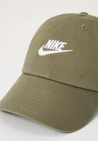 Alyvuogių žalia baseball tipo kepurė, pagaminta iš medvilnės, su baltu siuvinėtų „Nike“ logotipu priekyje, tekstūruotu paviršiumi ir išlenktu kraštu.