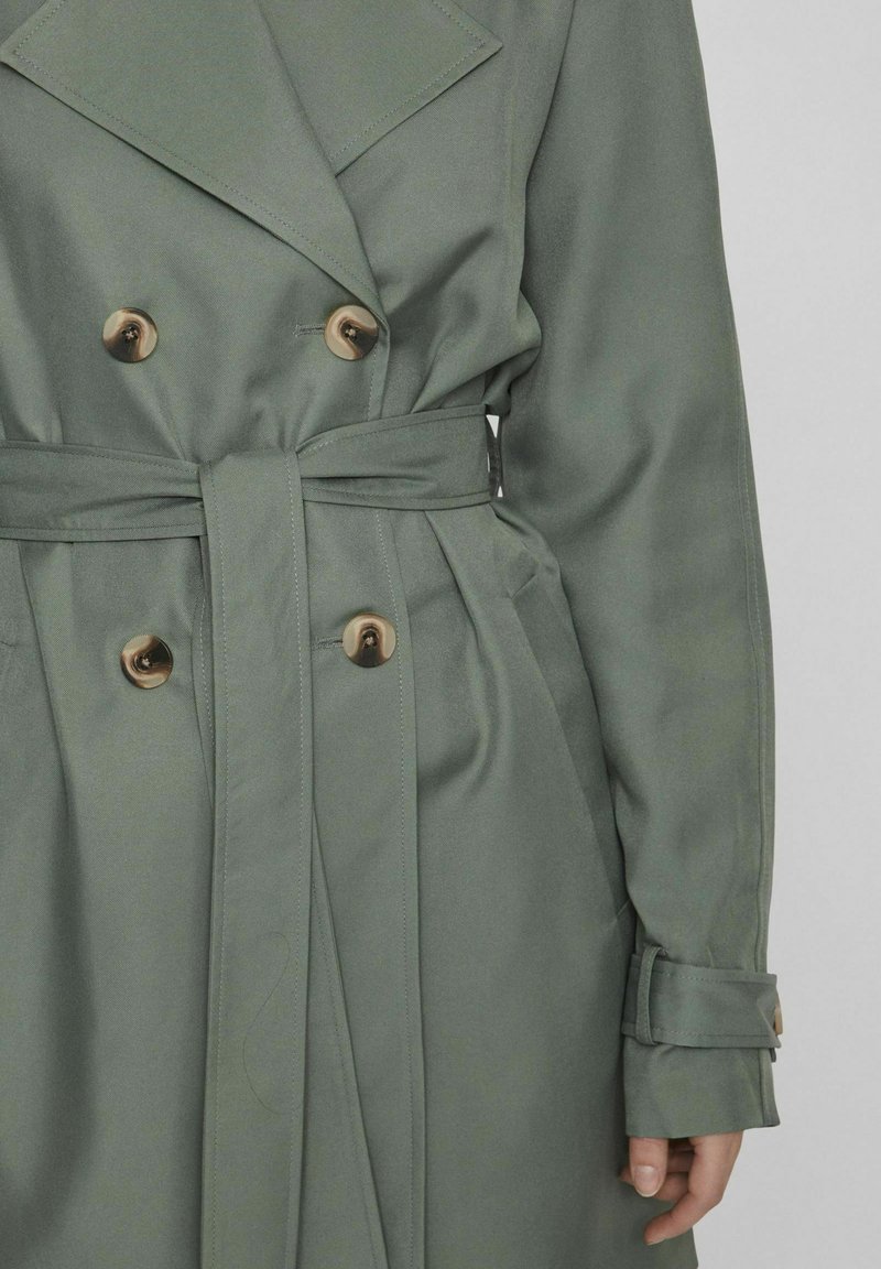 Dubbelgevoerde trenchcoat van olijfgroen stof, met grote beige knopen, een ceintuur in de taille en brede revers met manchetaccenten.