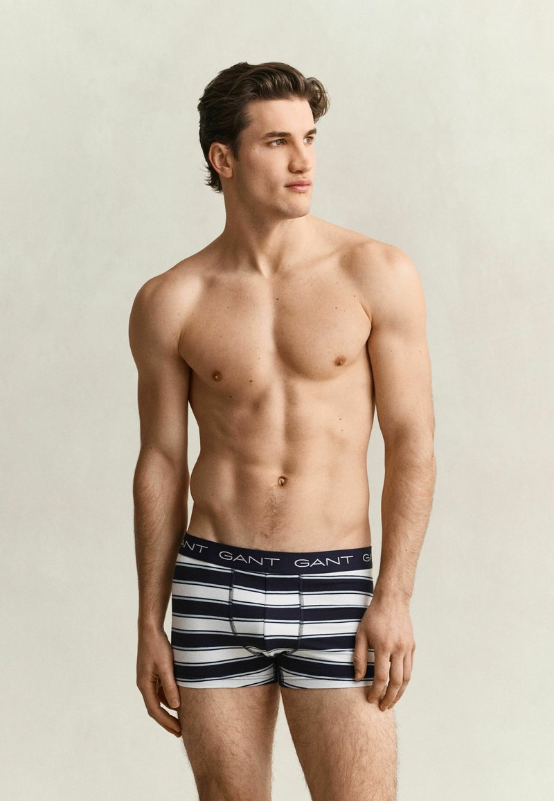Homme torse nu portant un boxer rayé noir et blanc avec le logo GANT, debout devant un fond clair uni, regardant vers la droite.