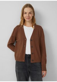 Brązowy sweter rozpinany z dekoltem w serek, sześcioma guzikami i ściągaczami na rękawach. Noszony na białej koszuli i zestawiony z ciemnymi dżinsami. Miękka faktura.