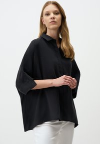 Black oversized shirt with a button-down collar, three-quarter sleeves, soft texture, and a curved hem, paired with white pants.
