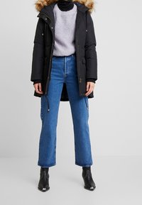 Parka acolchada negra con capucha de piel sintética, combinada con un suéter lila claro y jeans de tiro alto azul; botas negras hasta el tobillo con detalles plateados.