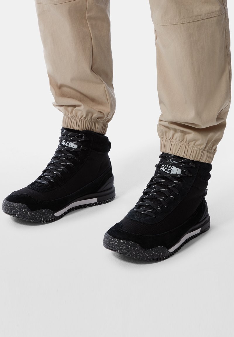 Baskets montantes noires avec semelles tachetées et lacets réfléchissants portées avec un pantalon beige retroussé sur fond blanc.