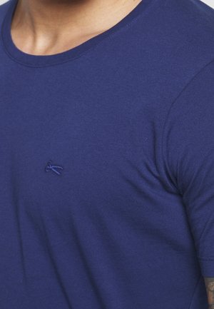 Nahaufnahme einer Person, die ein schlichtes marineblaues T-Shirt mit einem kleinen blauen gestickten Logo auf der linken Brust trägt.