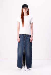T-shirt bianco a maniche corte, gonna lunga in denim blu scuro con spacco frontale e sneakers bianche. Un berretto nero completa l'outfit.