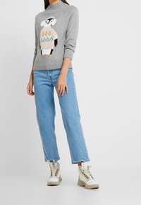 Grå stickad tröja med en pingvin-design och pastellfärgade accenter, som är kombinerad med ljusblå vida jeans och beige snörstövlar.