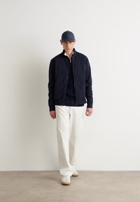 Navy bomber jacket with zip closure, paired with a dark sweater and white trousers. Complete with white sneakers and a matching cap.