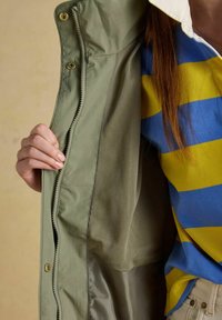 Mână care ține deschisă o jachetă verde măsliniu, arătând căptușeala din plasă, lângă un trunchi ce poartă un tricou cu dungi albastre și galbene și pantaloni bej.