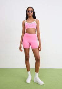 Ružový športový crop top a zodpovedajúce šortky s vysokým pásom, vyrobené z elastického materiálu s kontrastnými bielymi lemami, nosené s bielymi teniskami.