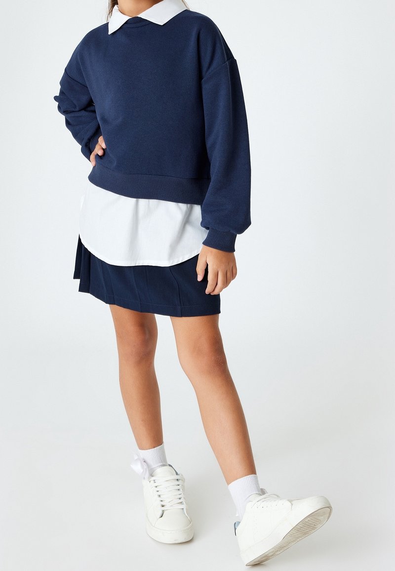 Enfant portant un pull bleu marine, une chemise blanche à col, une jupe bleu marine, des chaussettes blanches et des baskets blanches, debout avec une main sur la hanche.