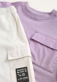 Fioletowa koszula z krótkim rękawem obok jasnokremowych spodni. Koszula ma kieszeń; spodnie posiadają markową etykietę i mankiety. Bawełniana tkanina, luźny krój.