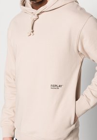 Uomo che indossa un hoodie beige chiaro con coulisse e il logo "REPLAY Second Life" sul davanti, con la mano nella tasca destra.