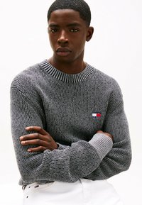 Maglione grigio a maglia con colletto e polsini a coste, caratterizzato da una piccola toppa con logo rosso, bianco e blu sul petto. Tessuto spesso, vestibilità morbida.