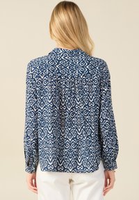 Blusa a righe blu navy e bianca con collo a camicia, polsini con bottone e dettaglio plissettato sul retro. Tessuto leggero e intrecciato con una consistenza morbida.