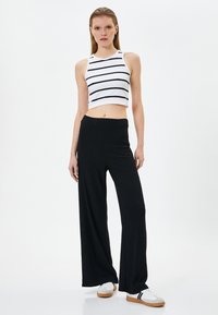 Zwarte wijde broek gecombineerd met een witte crop top met zwarte horizontale strepen. Model staat tegen een eenvoudige witte achtergrond en draagt sneakers.