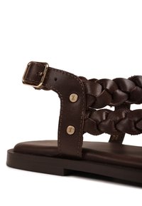 Donkere bruine leren sandaal met gevlochten bandjes, gouden gesp, twee decoratieve klinknagels en platte zool, getoond in een close-up zijaanzicht.