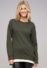 Pull avec manches longues couleur vert olive, à la texture douce, col rond et fentes sur les côtés. Design superposé avec un bas blanc visible.