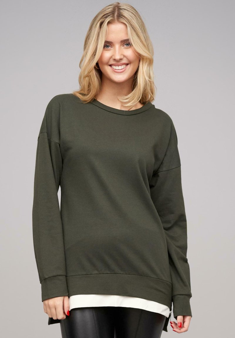 Pull avec manches longues couleur vert olive, à la texture douce, col rond et fentes sur les côtés. Design superposé avec un bas blanc visible.