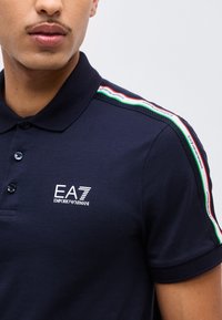 Κοντινό πλάνο ενός ατόμου που φοράει μπλε σκούρο πόλο με το λογότυπο EA7 Emporio Armani στο στήθος και ριγέ εμπριμέ κόκκινες, λευκές και πράσινες ρίγες στους ώμους.