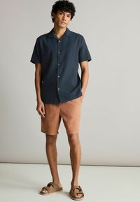 Camisa de botones de manga corta, de color azul oscuro, con un patrón texturizado, combinada con pantalones cortos de color marrón claro. El modelo lleva sandalias deslizantes marrones.