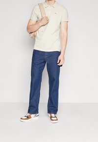 Beige Polo-Shirt, blaue gerade geschnittene Jeans und weiße Sneakers mit braunen und marineblauen Akzenten. Modell hält einen hellbeigen Rucksack.