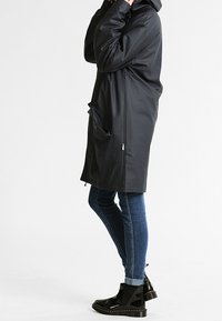 Czarny wodoodporny płaszcz z kapturem, bocznymi kieszeniami i luźnym krojem, noszony na niebieskich dżinsach i czarnych botkach z połyskującym wykończeniem.