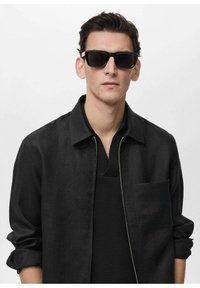 Black sunglasses with a rectangular frame paired with a black zip-front jacket and a textured black undershirt. Smooth finish, minimal design.