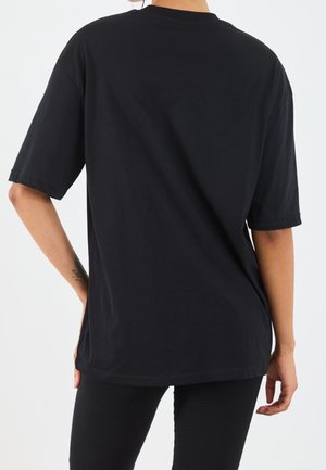 Osoba pokazana od tyłu ubrana w gładki, czarny, oversize t-shirt z krótkim rękawem i czarne spodnie na jednolitym, białym tle.