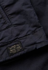 Ciemnogranatowy materiał o gładkiej fakturze. Widoczna jest czarna etykieta z złotym napisem „Superdry” oraz „Workwear”.