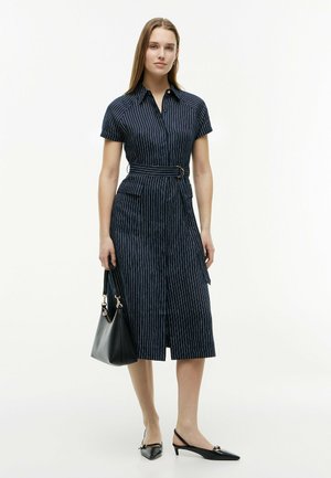 Frau trägt ein marineblau gestreiftes Kleid mit Gürtel, hält eine schwarze Handtasche und trägt schwarze Slingback-Pumps, steht vor weißem Hintergrund.
