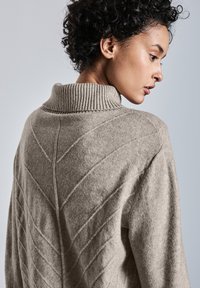 Maglione lavorato a maglia beige con colletto alto a coste, caratterizzato da un motivo a punto geometrico sul retro. Il tessuto appare morbido e con una texture interessante.
