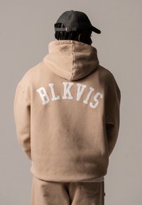Hanorac bej cu text alb "BLKVIS" în arc pe spate, având o glugă mare și o textură moale. Purtat cu o pălărie închisă la culoare.