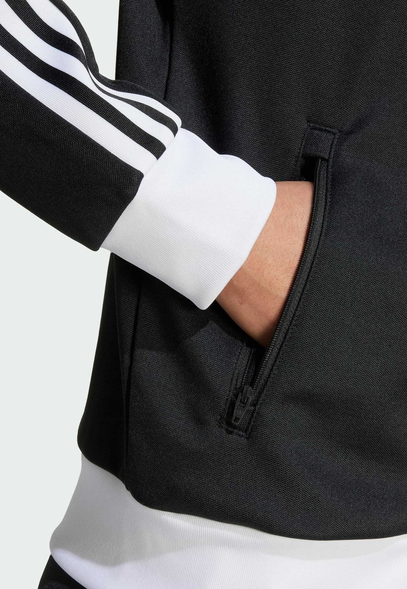 Sudadera deportiva negra con puños blancos y tres rayas verticales. Incluye un bolsillo con cremallera en el lateral con textura suave.