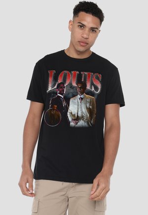 Junger Mann trägt ein schwarzes T-Shirt mit einer Grafik von drei Männern in Vintage-Anzügen und großem roten Schriftzug "LOUIS" auf der Vorderseite.