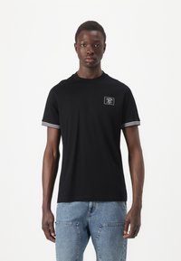 T-shirt preta de mangas curtas, com punhos listrados em branco, decote redondo e um pequeno rótulo frontal com detalhes em texto.