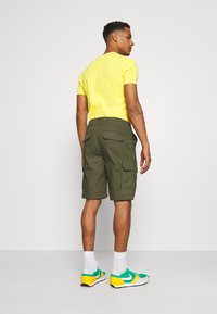 Olivenfargede cargo-shorts med baklommer, kombinert med en lys gul t-skjorte og multikolor sneakers. Enkelt bakgrunn.