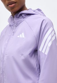 Personne portant une veste à capuche violet clair avec un logo blanc à trois bandes sur la poitrine et la manche, fermeture éclair à l'avant, vue en gros plan.