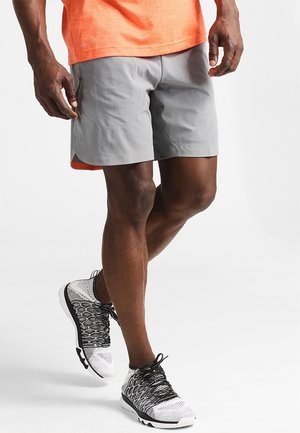 Mann trägt graue Sportshorts, oranges Shirt und schwarz-weiße Laufschuhe, steht mit einem Bein über das andere geschlagen.