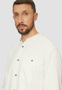Witte langemouwen shirt van een gestructureerde stof, met een bandkraag en twee borstzakken. Voorzien van donkere knopen en een relaxte pasvorm.