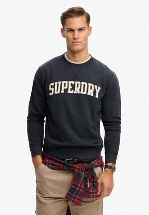 Uomo che indossa una felpa blu scuro con logo "SUPERDRY", pantaloni beige, camicia a quadri rossi e verdi legata in vita e un orologio da polso, in piedi con le mani nelle tasche.