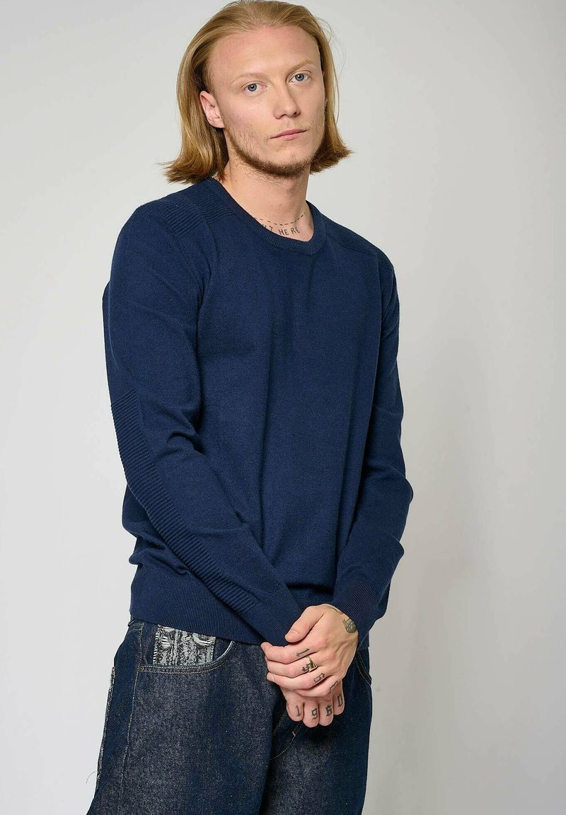 Pull en tricot bleu marine avec des accents côtelés sur les épaules et les poignets. Col rond, design ajusté et texture douce. Porté avec un pantalon en denim foncé.