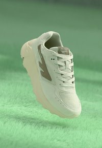 White athletic shoe with mesh fabric, textured accents in light brown, rounded shape, and thick sole, suspended against a green background.