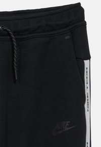 Μαύρο αθλητικό παντελόνι με κορδόνι στη μέση, μαλακή υφή, πλαϊνές τσέπες και την επιγραφή «Nike Tech» σε λευκό κατά μήκος της πλαϊνής ραφής.