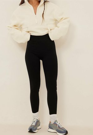 Vrouw draagt een crèmekleurige fleece trui met ritssluiting, zwarte hooggetailleerde legging, witte sokken en blauwgrijze sneakers, staand met handen in de zakken.