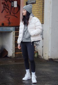 White puffer jacket with a high collar, gray hooded sweatshirt beneath, black leggings, and white sneakers. Gray beanie on head.