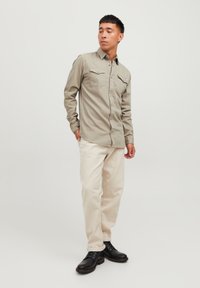 Camisa de botón de un verde oliva claro con dos bolsillos en el pecho, combinada con pantalones color crema y zapatos negros, sobre un fondo claro.