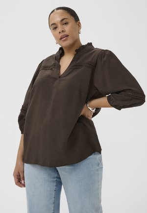Femme portant un chemisier ample marron foncé avec des détails en dentelle et un jean bleu clair, posant avec une main sur la hanche contre un fond uni.