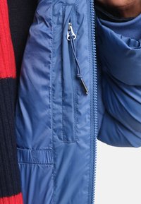 Modrá vycpaná bunda s bližším zapínáním, elastickým detailem a texturovanými akcenty. Hladký vnější materiál s teplou podšívkou.